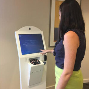 A woman with dark hair wearing a navy blue top and lime green skirt uses a patient check-in kiosk. Her right hand is extended to touch the screen, and a scanner or payment slot is visible below the screen. A framed picture hangs on the wall in the background.