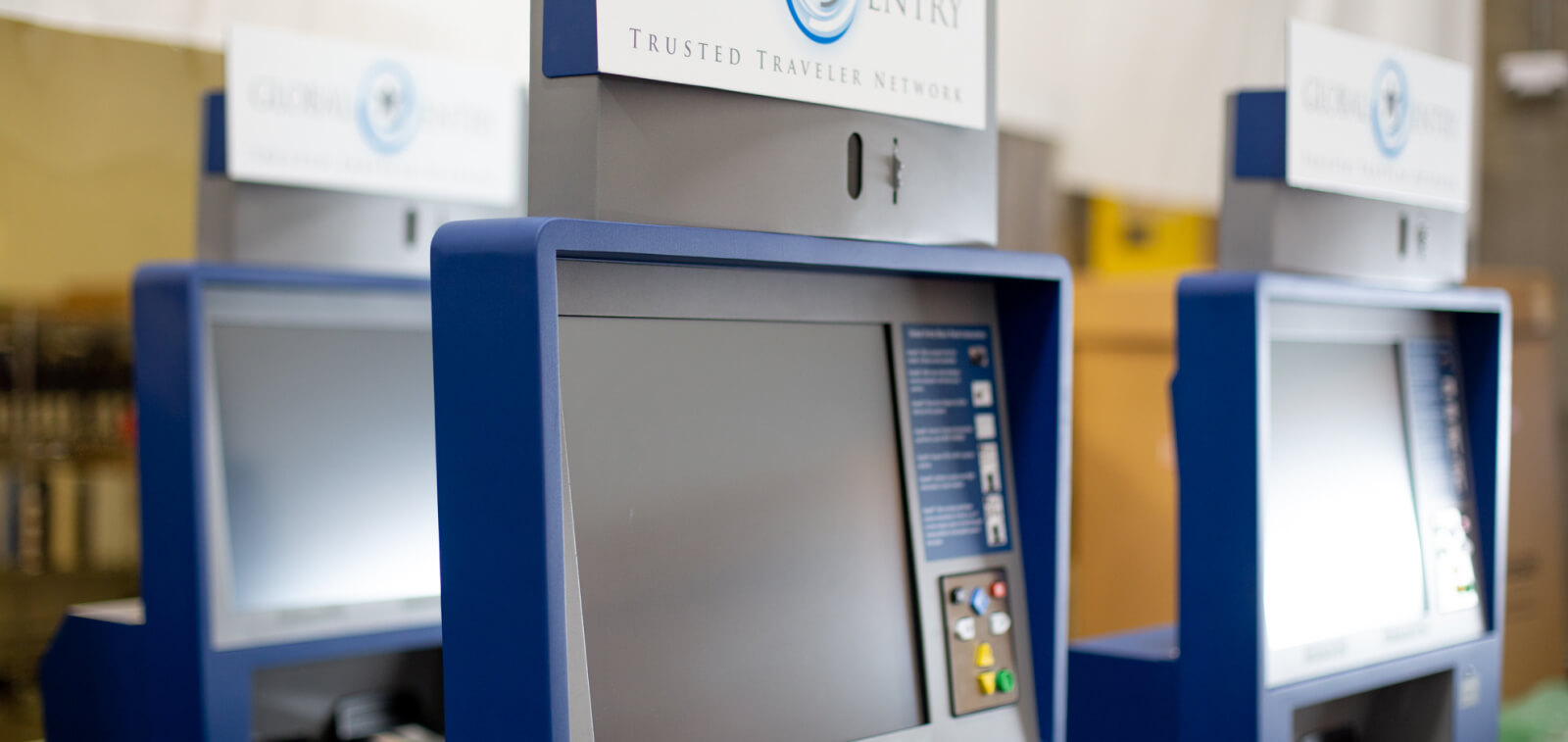 Close up of Global Entry Kiosk, in a warehouse facility. Kiosk is blue and grey with touch screen capability