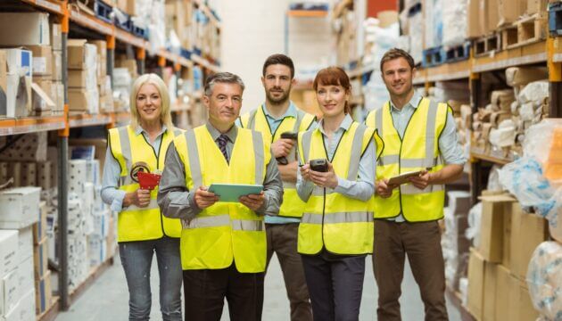 Smiling warehouse team looking at camera, wearing yellow saftey vests and holding equipment