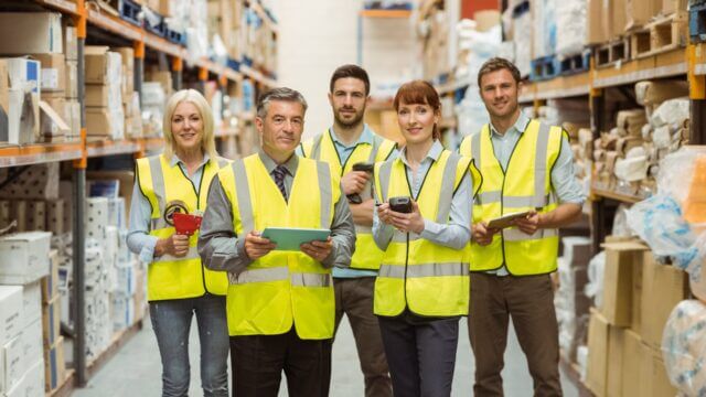 Smiling warehouse team looking at camera, wearing yellow saftey vests and holding equipment