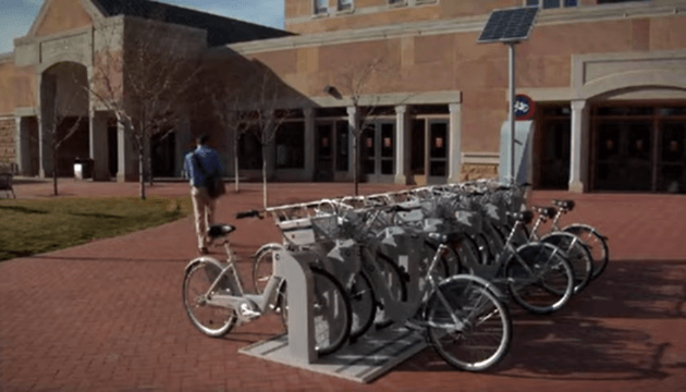 Row of rental bicycles in an outdoor setting. They are attached a rental kiosk that holds them in place by their front wheel