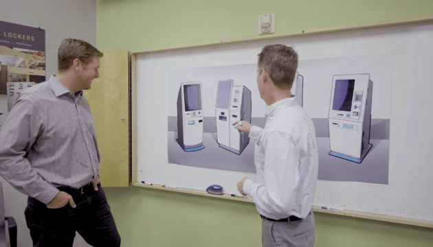 Two men in an office setting looking at drawings of kiosks on a board