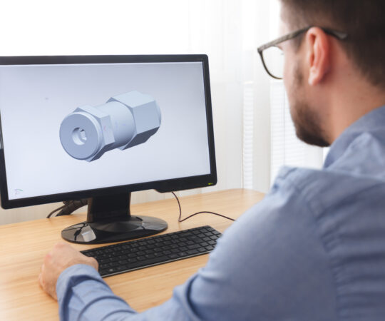 Over-the-shoulder of man sitting at a desk, using a desktop computer. He is working on 3D rendering.