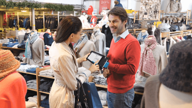 A man and woman instead a bright department store. The woman is using a handheld kiosk device to pay for something