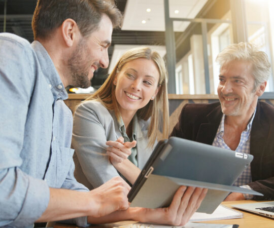 Three people sitting at a table, smiling and looking at an ipad monitor