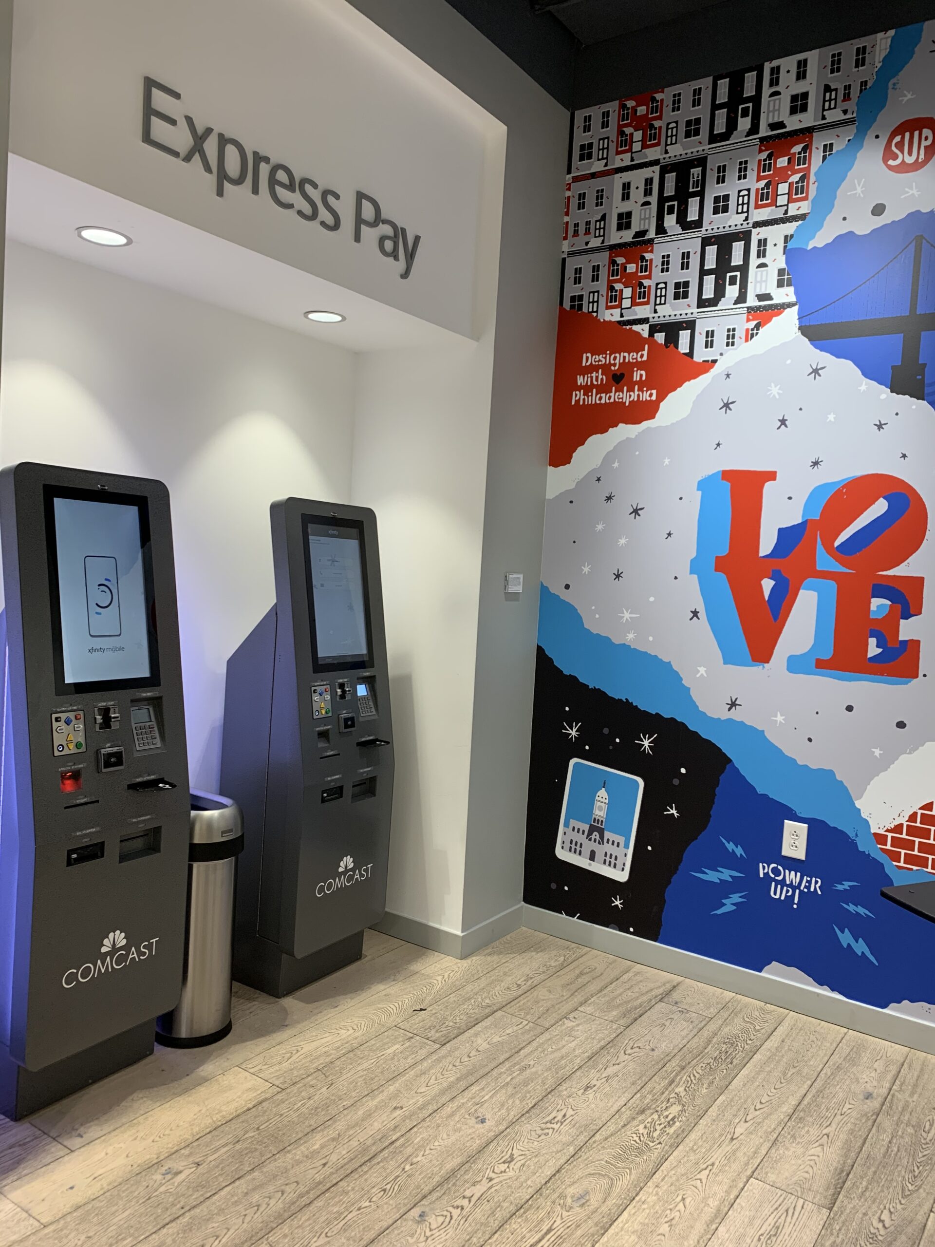 Inside a Comcast store, showcasing two grey bill payment kiosks. They have large touchscreens and multiple keypads for payment options