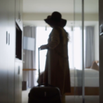 Woman wearing a hat walking into a hotel room with a rolling suitcase