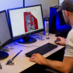 Man sitting at desk working with two desktop monitors. He is working on a 3D rendering of a red kiosk