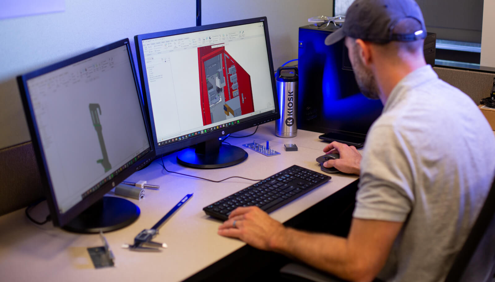 Man sitting at desk working with two desktop monitors. He is working on a 3D rendering of a red kiosk