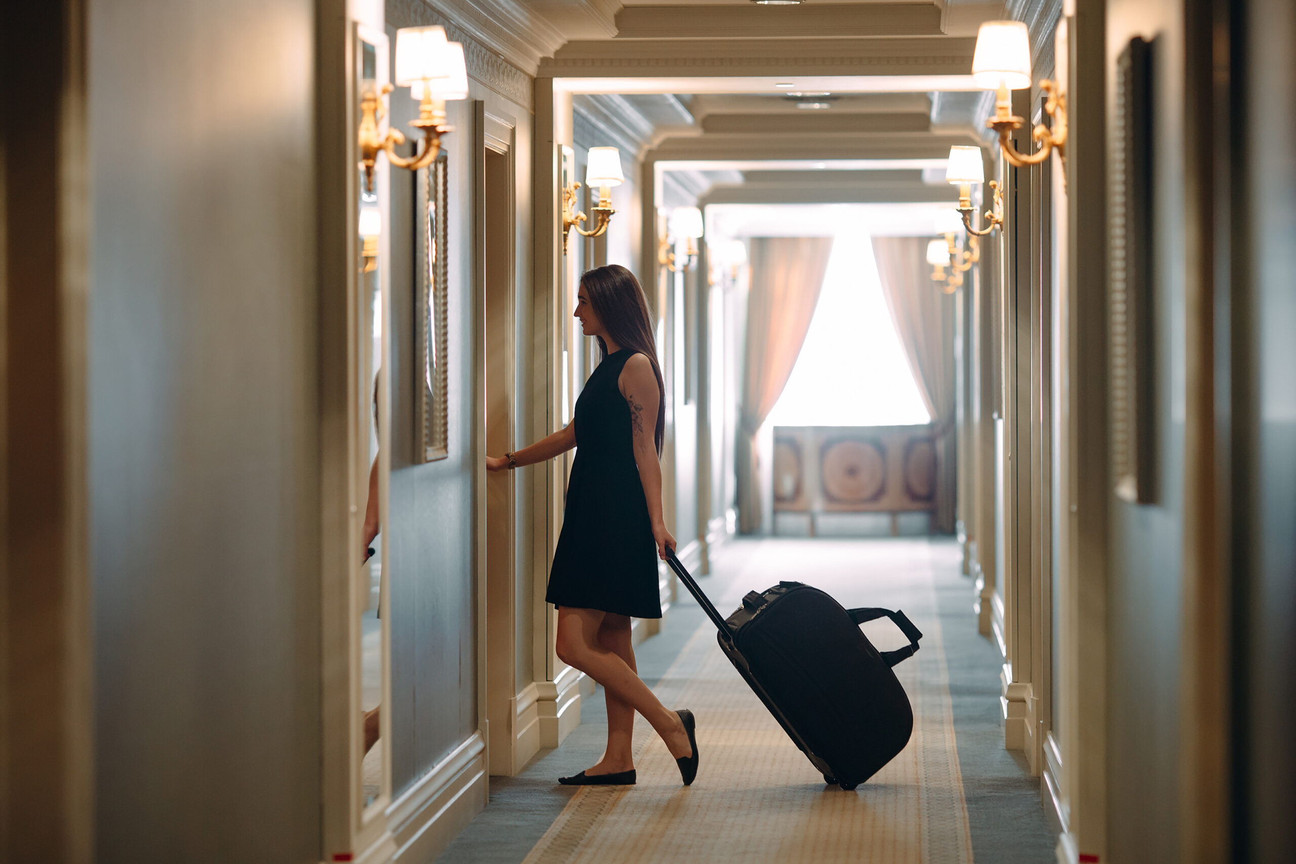 Young woman with handbag and suitcase in an elegant suit walks the hotel corridor to her room